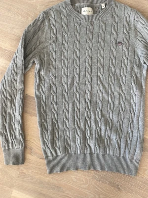 Grå kabelstickad tröja från GANT - Snygg grå kabelstickad tröja från GANT med rund halsringning och ribbade muddar. Tröjan har ett broderat GANT-emblem på bröstet och är tillverkad i mjukt material. Perfekt för dig som gillar klassisk och stilren look. Fint skick. Stl xs