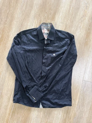 Svart skjorta från Burberry - Stilren svart skjorta från Burberry med klassiskt rutigt mönster på insidan av kragen och knappslån. Skjortan har långa ärmar, knappar framtill och en liten logga på bröstet. Perfekt för dig som gillar exklusiva detaljer och tidlös design.
