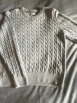 Helt oanvänd Ljusgrå kabelstickad tröja från Gant  - Kabelstickad tröja från Gant i ljusgrå färg med rund halsringning och ribbade muddar. Klassisk design med diskret Gant-logga på bröstet. Perfekt för dig som gillar stilrena och tidlösa plagg.