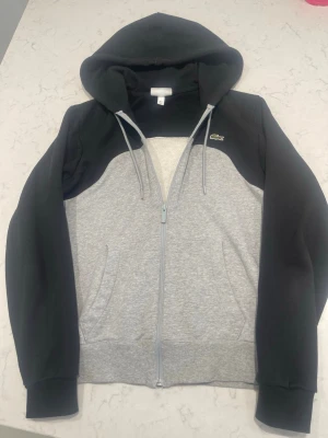 Grå och svart hoodie från Lacoste - Snygg hoodie från Lacoste i klassisk fit med dragkedja framtill. Tröjan är grå på kroppen och svart på ärmar och huva. Har två fickor fram och den ikoniska gröna krokodilloggan på bröstet. Tillverkad i mjukt bomullsmaterial och har justerbar huva med snören. Sjukt fet kofta nu till sommaren och nypris ligger på ca 1600kr