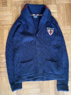 Mörkblå cardigan från Polo Ralph Lauren - Mörkblå kabelstickad kofta med sjalkrage och knappar framtill. Två fickor fram och ett broderat märke på bröstet. Kragen har en röd detalj på insidan. Perfekt för dig som gillar klassisk och stilren look.
