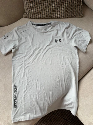 Under Armour UA-SPEC träningsshirt – grå – storlek M - Ljusgrå tränings-/löpartröja från Under Armour i storlek M. UA-SPEC-design med svarta logodetaljer framtill och längs sidan. Lätt och funktionellt material som passar bra för löpning, gym och annan träning.  Fint begagnat skick.