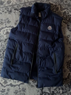 Mörkblå dunväst från Moncler - Snygg mörkblå dunväst från Moncler med klassisk logga på bröstet. Västen har två dragkedjefickor framtill och hög krage. Perfekt för lager-på-lager och ger en sportig vibe. Fodret har en unik serietidningsetikett på insidan.