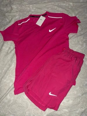Nike miler - Snyggt rosa träningsset från Nike med t-shirt och shorts. T-shirten har vita detaljer och Nike-logga på bröstet, medan shortsen har resår i midjan och vit logga på benet. Perfekt för löpning eller gym. Materialet är lätt och andas.