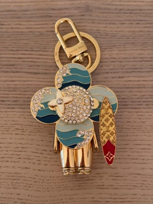 Vuitton surf-berlock i guld - Unik accessoar från Vuitton i form av en surfande figur. Tillverkad i guldfärgad metall med blå, vita och turkosa vågmönster samt gnistrande stenar. Surfbrädan har röda och guldiga detaljer med LV-monogram. Perfekt som nyckelring eller väsksmycke.
