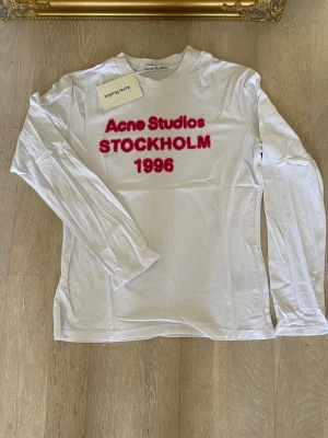 Vit långärmad tröja Acne Studios M - Säljer en vit långärmad topp från Acne Studios med tryck i rött framtill där det står 'Acne Studios STOCKHOLM 1996'. Tröjan har rund halsringning och är gjord i ett tunt, mjukt material som känns skönt mot huden. Perfekt för en trendig och avslappnad stil.