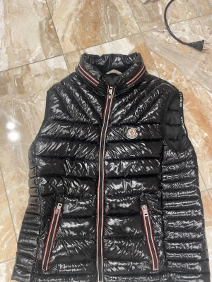 Svart dunväst från Moncler - Snygg svart dunväst från Moncler med glansigt material och quiltad design. Västen har ståkrage, två fickor med dragkedja och Moncler-logga på bröstet. Dragkedjorna har rödvita detaljer som ger en sportig touch. Perfekt för lager-på-lager-stil.