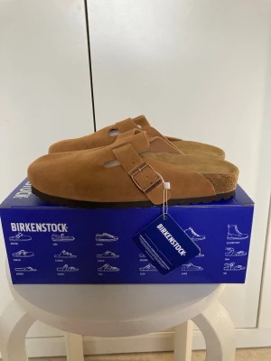 Bruna Birkenstock Boston tofflor - Säljer ett par klassiska Birkenstock Boston tofflor i brun mocka med justerbart spänne och bekväm korksula. Perfekta för dig som vill ha både stil och komfort hemma eller ute. Tidlös design och riktigt sköna att gå i.