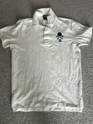 Vit Ralph Lauren Rugby t-shirt - Vit t-shirt från Ralph Lauren Rugby med klassisk krage och korta ärmar. På bröstet finns en mörkblå döskalle-brodyr som ger en cool touch. Tillverkad i mjuk bomull som känns skön mot huden. Perfekt för dig som gillar stilrena plagg med en twist. Denna är M i storlek men har krymp lite! Använd ganska mycket så helt ok skick! Säljer för 400 kr för denna är väldigt svår att få tag på och väldigt unik 