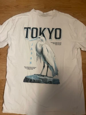 Vit T-shirt med Tokyo-tryck - Vit T-shirt med stort tryck på ryggen som föreställer en häger och texten 'TOKYO JAPAN' i blått och svart. Framsidan har en mindre Tokyo-text på bröstet. Klassisk passform och rund hals. Perfekt för dig som gillar grafiska motiv och streetwear.