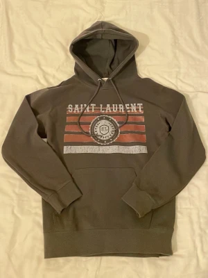 Grå Saint Laurent hoodie med tryck - Snygg grå hoodie från Saint Laurent med huva och dragsnören. Framtill finns ett tryck med texten 'SAINT LAURENT' och röda samt vita ränder, samt ett emblem i mitten. Hoodien har ribbade muddar vid ärmar och nederkant samt en klassisk magficka.