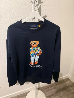 Mörkblå sweatshirt Polo Bear - Mörkblå sweatshirt från Polo Ralph Lauren med ikoniska Polo Bear-trycket framtill. Björnen har på sig en färgglad skjorta, vita byxor och solglasögon. Tröjan har rund halsringning och ribbade muddar. Perfekt för en avslappnad och trendig stil. Säljer den då den va för stor för mig. Den är alltså helt ny.
