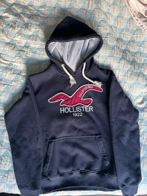 Mörkblå Hollister hoodie med logga - Snygg mörkblå hoodie från Hollister med stor broderad logga och texten 'California 1922' på bröstet. Klassisk känguruficka framtill, vit dragsko i huvan och mjukt innertyg. Perfekt för chill dagar eller när du vill ha en avslappnad stil.