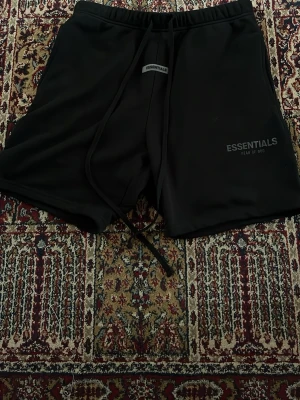 Essentials Fear of God - Svarta shorts från Essentials Fear of God med snörning i midjan och logga tryckt på vänster ben. Klassisk passform och mjukt material som känns skönt mot huden. Perfekta för en avslappnad streetwear-look.