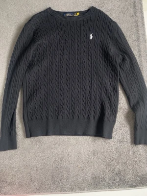 Mörkgrå kabelstickad tröja Polo Ralph Lauren - Snygg mörkgrå kabelstickad tröja från Polo Ralph Lauren med klassisk vit logga på bröstet. Tröjan har rund halsringning, långa ärmar och ribbade muddar. Perfekt för dig som gillar stilrena och tidlösa plagg.