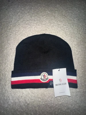 Svart Moncler mössa med ränder - Snygg svart mössa från Moncler med vikbar kant och klassiska Moncler-loggan framtill. Mössan har blå, vit och röd rand längs kanten för en sportig vibe. Tillverkad i mjukt, ribbstickat material som sitter skönt på huvudet. Perfekt accessoar för kalla dagar.