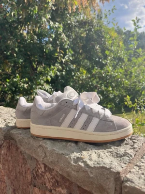 Grå Adidas Campus sneakers - Snygga grå Adidas Campus sneakers i mocka med vita detaljer och klassiska tre ränder på sidan. Skorna har vit sula och breda vita skosnören. Perfekta för dig som gillar retrostil och vill ha något bekvämt och stilrent till vardags.