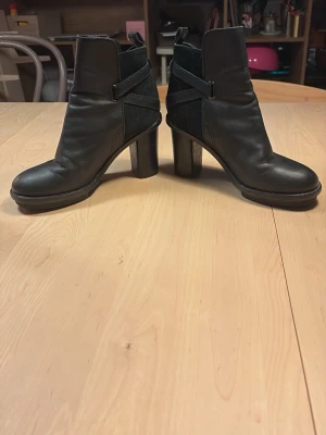 Svarta stövletter med klack - Snygga svarta stövletter i skinn med rund tå och bred klack. Dekorativa remmar runt ankeln och detaljer i mocka baktill ger en cool look. Perfekta till jeans eller kjol för en trendig outfit.
