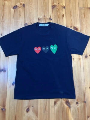 Mörkblå Comme des Garçons PLAY t-shirt - Säljer en mörkblå t-shirt från Comme des Garçons PLAY med tre hjärtan i rött, svart och grönt på bröstet. Klassisk rund hals och korta ärmar. Perfekt för dig som gillar streetwear och vill sticka ut lite extra.