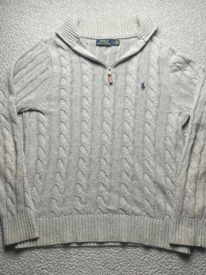 Grå kabelstickad tröja Polo Ralph Lauren - Säljer en grå kabelstickad tröja från Polo Ralph Lauren med halvzip och hög krage. Mycket bra skick, skickar snabbt efter att du har beställt.
