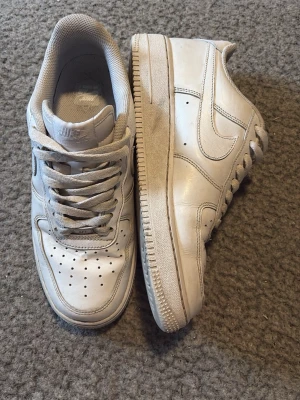 Nike Air Force 1 vita sneakers - Klassiska Nike Air Force 1 sneakers i helvitt läder med perforerad tå, vita skosnören och den ikoniska swoosh-loggan på sidan. Tjock sula och låg profil ger en tidlös look som alltid funkar. Perfekta för dig som gillar streetwear och enkel stil. Dom är använda men går jätte fort att få dom att se hel nya ut. Hör av vid frågor!