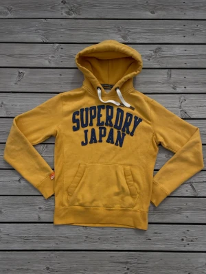 Superdry hoodie - Gul hoodie från Superdry med stor blå text 'SUPERDRY JAPAN' på bröstet. Har inte hittat någon exakt likadan hoodie och tror därför den är sällsynt, så var snabb!! Fråga gärna vid intresse, priset alltid diskuterbart😉