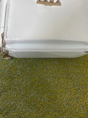 Vit handväska från Zara - Snygg vit handväska från Zara i slätt konstläder med guldfärgade detaljer och dragkedjor. Väskan har en boxig form och justerbar axelrem. Perfekt för dig som vill ha en stilren och trendig accessoar.