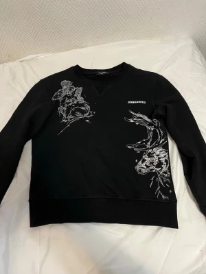 Svart sweatshirt från Dsquared2 - Säljer en svart sweatshirt från Dsquared2 med vita broderade djurmotiv på framsidan. Tröjan har rund halsringning och ribbade muddar vid ärmslut och nederkant. Stilen är clean men ändå unik tack vare de detaljerade broderierna och den diskreta loggan på bröstet.