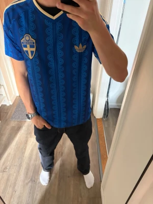 Blå Adidas Sverige fotbollströja - Säljer en blå fotbollströja från Adidas med Sveriges landslagsemblem på bröstet och guldfärgade detaljer. Tröjan har ett mönster i tyget, korta ärmar och klassiska Adidas-ränder på axlarna. Perfekt för match eller träning!