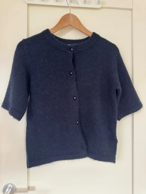 Mörkblå stickad kofta Gina Tricot - Mysig mörkblå stickad kofta från Gina Tricot med korta ärmar och rund halsringning. Koftan har knappar framtill och är gjord i ett fluffigt material som ger en soft känsla. Perfekt att slänga över en topp för en chill look.