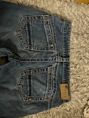 Blå bootcut jeans med kontrastsömmar - Snygga blå bootcut jeans med tydliga kontrastsömmar och stora bakfickor. Jeansen har en klassisk femficksmodell och en liten läderpatch bak i midjan. Perfekta för dig som gillar en avslappnad men trendig stil. Storlek 27/32, nypris-799