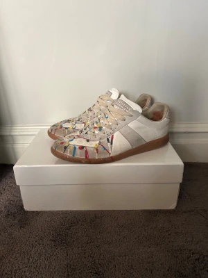 Maison Margiela Gats färgstänk - Maison Margiela Gats skor med coola färgstänk i blått, rött, gult och svart. Skorna har en klassisk låg profil och bruna sulor. Materialet är en mix av mocka och läder, vilket ger en snygg och trendig look. Perfekt för dig som vill sticka ut med dina skor.