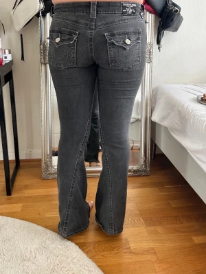 Grå bootcut jeans True Religion - PRIS GÅR ATT DISKUTERA!! Säljer ett par grå bootcut jeans från True Religion med dekorativa sömmar och fickor bak med lock och knappar. Jeansen har normal midja och klassisk femficksmodell. Snygg tvätt och coola detaljer som ger en trendig look.