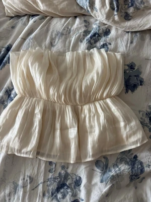 Off shoulder plisserad topp i creme - Säljer en superfin axelbandslös topp i cremefärgat, lätt och plisserat tyg. Toppen har en volangig, luftig passform och markerad midja som ger en romantisk vibe. Perfekt till jeans eller kjol för en trendig look.