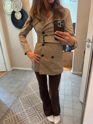Beige trenchcoat med bälte från J&J - Stilren beige trenchcoat från J&J med dubbelknäppning, axelklaffar och avtagbart bälte i midjan. Jackan har klassisk krage, sidofickor och dekorativa knappar. Perfekt för vår och höst, tillverkad i ett lätt tyg som ger en snygg siluett. Jättefint skick.
