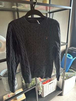 Mörkblå kabelstickad tröja GANT - Kabelstickad mörkblå tröja från GANT med rund halsringning och broderad logga på bröstet. Tröjan har långa ärmar och ribbade muddar vid ärmslut och nederkant. Perfekt för dig som gillar klassisk och stilren look.