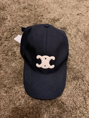 Celine keps med Triomphe-logo - Snygg svart keps från Celine med den ikoniska Triomphe-loggan broderad framtill i vitt. Kepsen är justerbar baktill och har Celine-tryck på insidan samt etikett kvar. Passar perfekt till både vardag och sportiga outfits.
