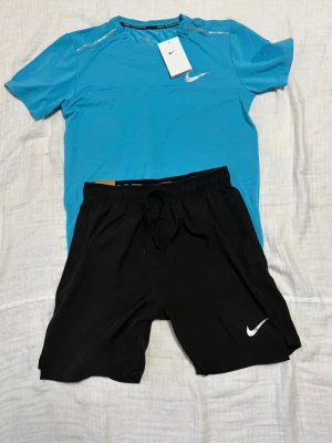 Nike träningsset blå t-shirt & svarta shorts - Nike träningsset med en blå Dri-FIT t-shirt och svarta shorts. T-shirten har silverfärgade detaljer och Nike-logga på bröstet, medan shortsen har en vit Nike-logga och dragsko i midjan. Perfekt för löpning eller gym. SITTER SOM STORLEK S ÄVEN FAST DE STÅR M