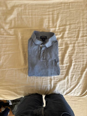 Blå piké t-shirt från Polo Ralph Lauren - Snygg blå piké t-shirt från Polo Ralph Lauren med klassisk krage och två knappar framtill. T-shirten har korta ärmar och den ikoniska broderade loggan på bröstet. Perfekt till jeans eller shorts för en clean och stilren look.