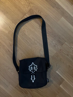 Svart axelväska från Trapstar - Snygg svart axelväska från Trapstar med justerbar axelrem och coolt vitt emblem på locket. Väskan har ett spänne framtill och meshficka på framsidan. Perfekt för att bära det viktigaste och ger en streetwear vibe.