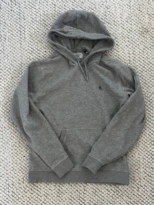 Grå hoodie från Penguin - Snygg grå hoodie från Penguin med klassisk huva och dragsko. Tröjan har en liten broderad logga på bröstet och en stor magficka framtill. Tillverkad i mjukt bomullsmaterial som känns skönt mot huden. Perfekt för en avslappnad och stilren look.