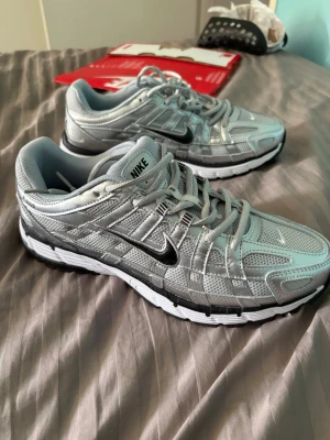 Nike silverfärgade mesh sneakers - Säljer ett par riktigt snygga Nike sneakers i silver och grått med svart swoosh. Skorna har en sportig design med mesh och syntetpaneler för bra andningsförmåga. Sulan är vit med svarta detaljer och ger grym komfort. Perfekta för dig som gillar streetwear.