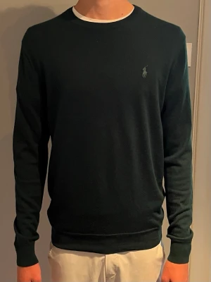 Mörkgrön tröja från Polo Ralph Lauren - Snygg mörkgrön tröja från Polo Ralph Lauren med diskret logga broderad på bröstet. Tröjan har rund halsringning, långa ärmar och ribbade muddar vid ärmslut och nederkant. Perfekt till jeans eller chinos. Nypris runt 2000kr. Sparsamt använd, killen på bilden är 190 och väger cirka 70 kg. 