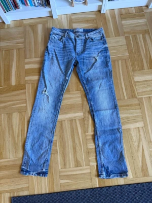 Ljusblå slitna jeans med hål - Säljer ett par ljusblå jeans med slitningar och hål på benen. Jeansen har klassisk femficksmodell, normal midja och raka ben. Materialet är denim i bomull och de har en avslappnad, trendig look.