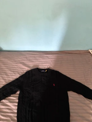 Svart kabelstickad tröja Polo Ralph Lauren - Kabelstickad svart tröja från Polo Ralph Lauren med rund halsringning och den klassiska röda logotypen broderad på bröstet. Tröjan har ribbade muddar vid ärmslut och nederkant. Perfekt för dig som gillar stilrena och tidlösa plagg.