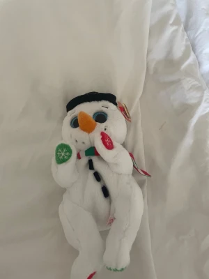 Mjuk snögubbe med färgglada vantar - Söt snögubbe i plysch med svart hatt, orange morotsnäsa och stora blå ögon. Har en randig halsduk i rött, grönt och vitt samt vantar i grönt och rött med snöflingemönster. Perfekt som juldekoration eller mysig leksak.