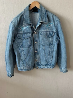 Roccobarocco Ljusblå jeansjacka med broderi - Snygg ljusblå jeansjacka från Roccobarocco med  unika blå broderier på bröstet och ryggen. Klassisk modell med knappar framtill, två bröstfickor med lock och dekorativa sömmar. Jackan har en avslappnad passform och coola detaljer längst ner.0069
