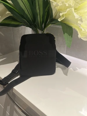 Svart axelväska från Hugo Boss sällsynt!! - Snygg svart axelväska från Hugo Boss med präglad logga på framsidan. Väskan har en fyrkantig form och justerbar axelrem. Tillverkad i ett slätt material som ger en stilren look. Perfekt för att bära det viktigaste med stil.