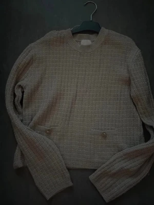 Beige stickad tröja med fickor - Mysig beige stickad tröja med rund halsringning och två små fickor framtill som har dekorativa knappar. Tröjan har lång ärm och ett diskret rutmönster i stickningen. Perfekt för lager på lager under kyliga dagar.