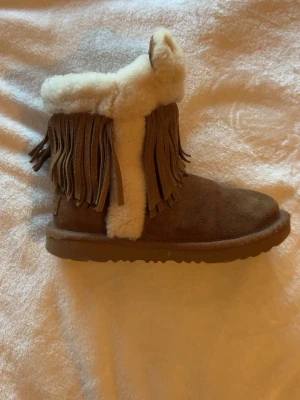 Bruna UGG boots | special edition 2024 - Säljer ett par bruna UGG boots i mocka med fluffig vit insida och dekorativa mockafransen längs skaftet. Skorna har en klassisk rund tå och grov sula, perfekta för kalla dagar. Mysig och trendig design som sticker ut.